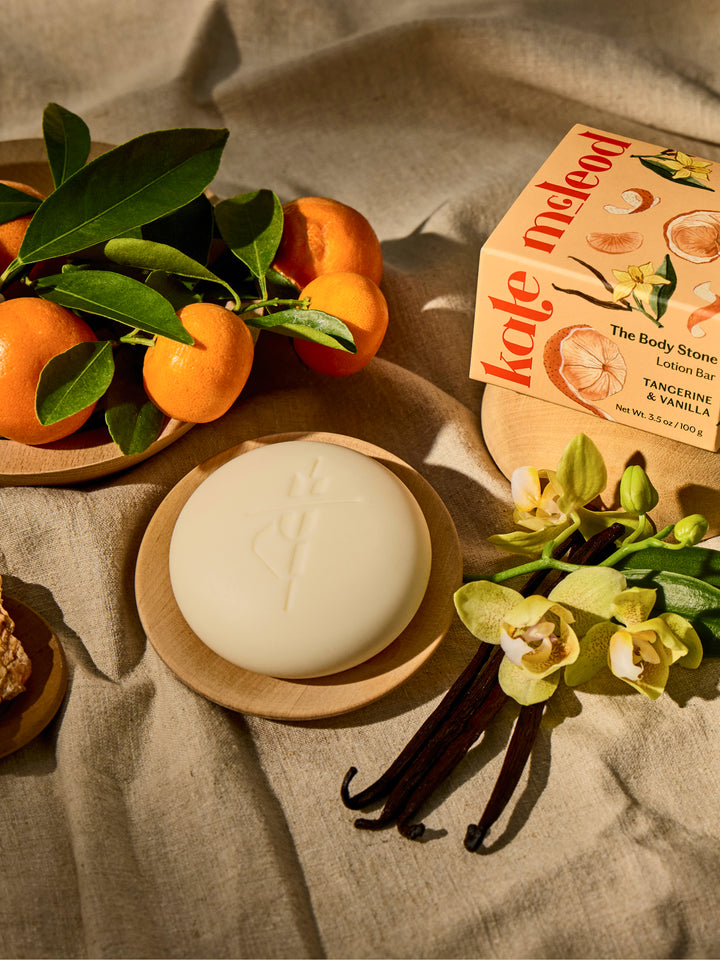 Kate McLeod Tangerine & Vanilla lotion bar with packaging on a wooden plate with ingredients and linens around