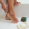 A person sitting on a white bathtub files the heal of their foot with the Kate McLeod foot file. The foot stone rest on the tub.