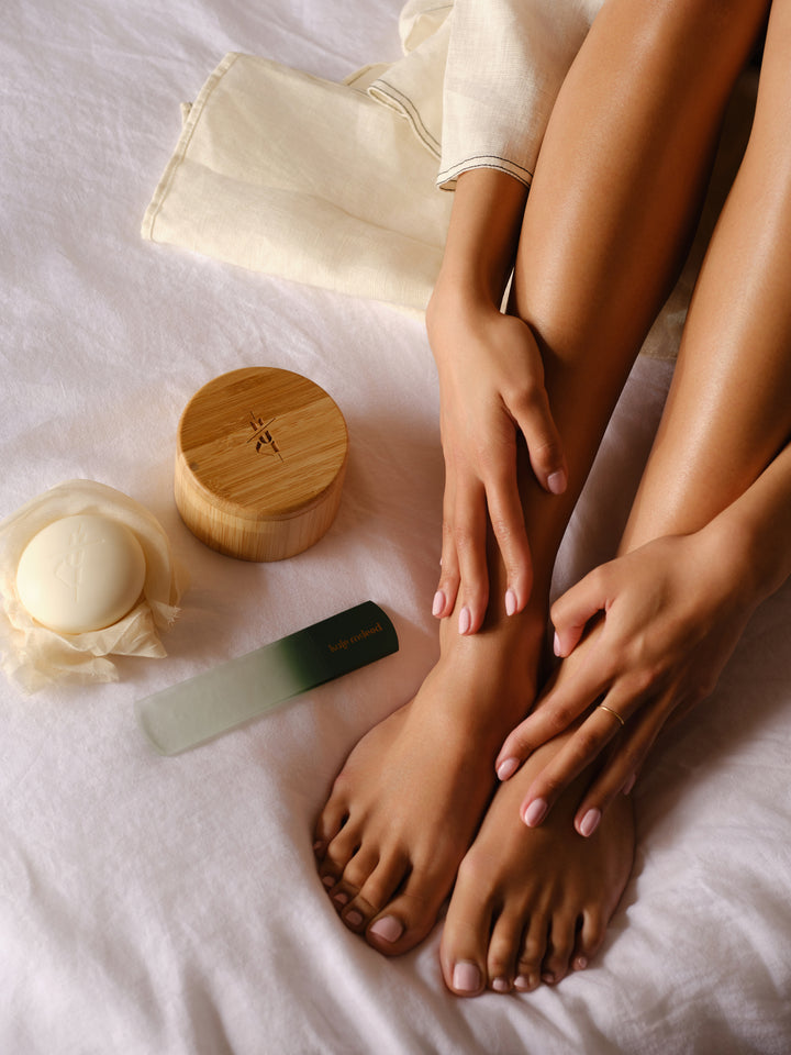 Person's legs and feet with the Kate McLeod reusable body stone canister, foot stone, and foot file on a white surface