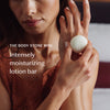 A woman holds a small white round lotion bar labeled 'THE BODY STONE MINI' against a softly blurred bedroom background. White text on the left reads 'Intensely moisturizing lotion bar.