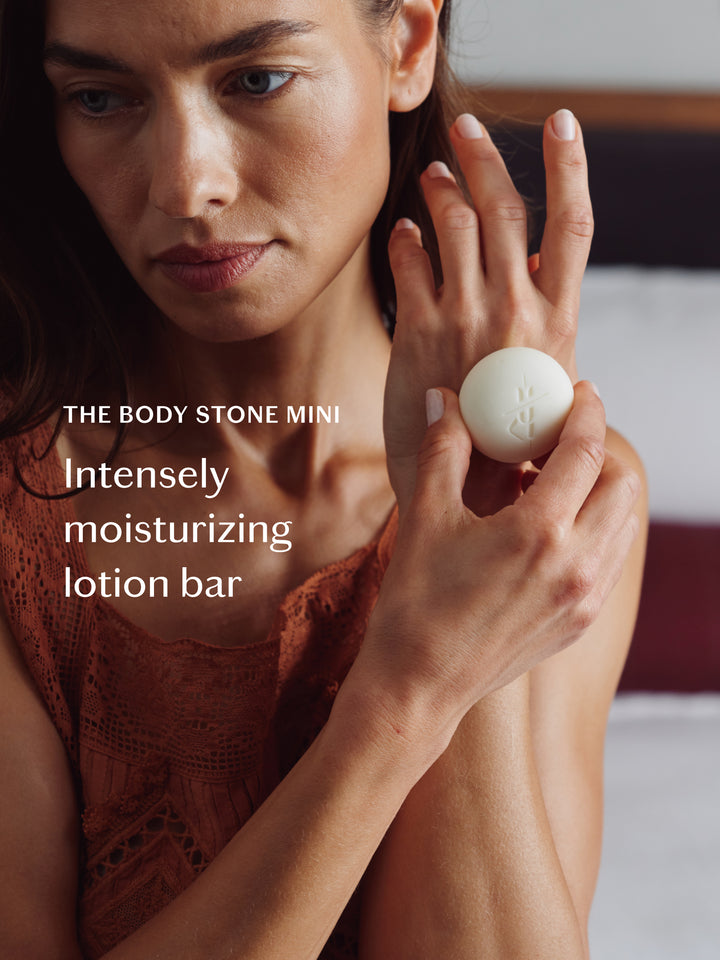 A woman holds a small white round lotion bar labeled 'THE BODY STONE MINI' against a softly blurred bedroom background. White text on the left reads 'Intensely moisturizing lotion bar.