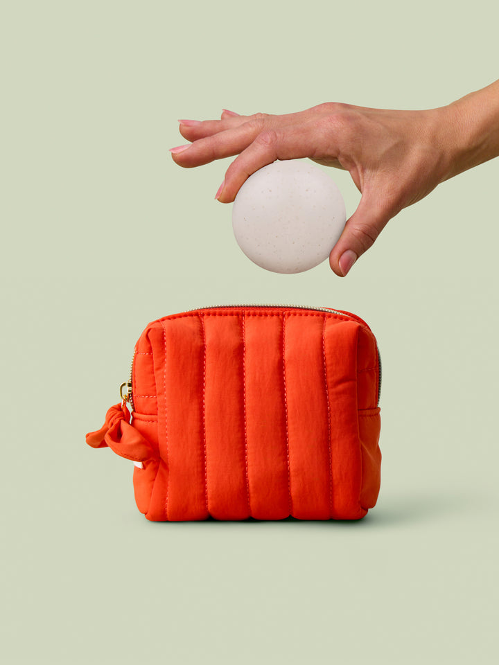 Hand holding a body stone above the poppy quilted mini travel bag on a light green background
