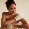 Woman exfoliating arms with the body mitt, sitting on a wooden bench.