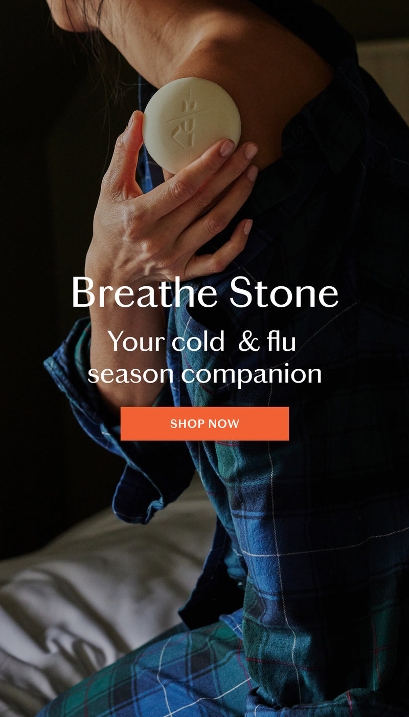 A person in a blue plaid shirt holds a round beige Breathe Stone against their neck. The image features promotional text reading 'Breathe Stone Your cold & flu season companion' with an orange 'SHOP NOW' button.