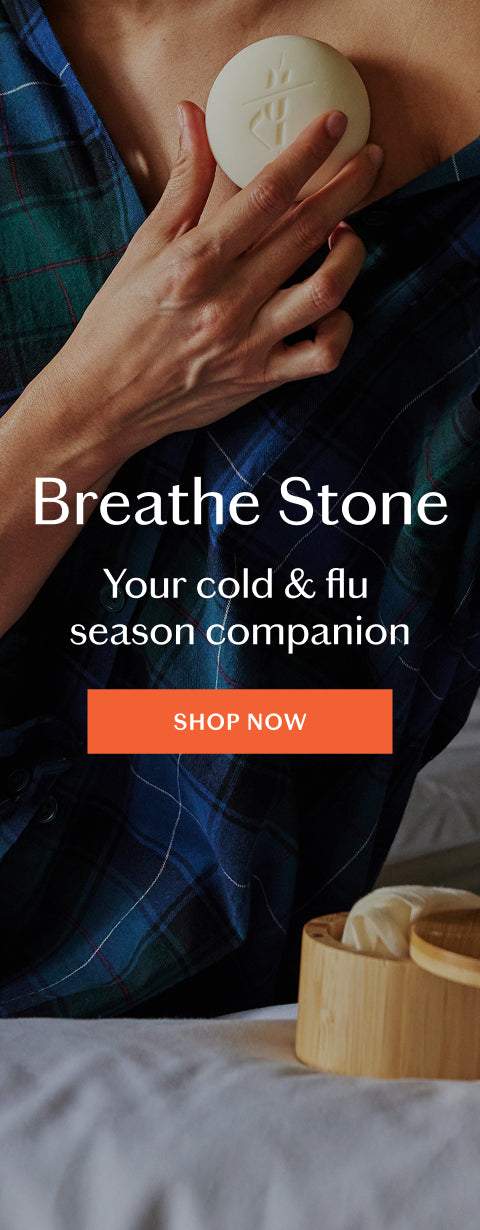 A person in a blue plaid shirt holds a round off-white stone near their chest, with overlaid text reading 'Breathe Stone' and 'Your cold & flu season companion' plus an orange 'SHOP NOW' button. A small bamboo container with more stones sits on a bed in the foreground.