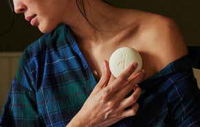 A close-up lifestyle shot of a woman wearing a blue and green plaid shirt, holding a round, breathe stone body stone against her chest. The soap features an embossed symbol resembling a stylized 'P' or medical caduceus mark.