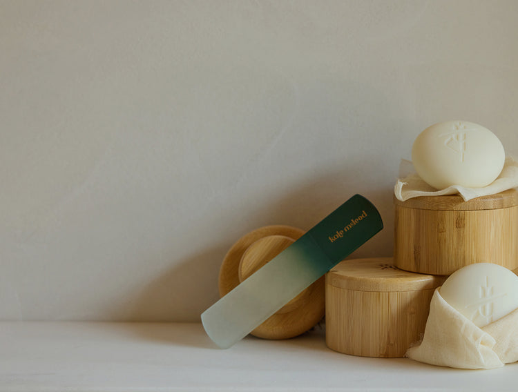 A foot file and gold 'Kate McLeod' branding, styled on a light wood surface next to bamboo containers and cream-colored body stones.