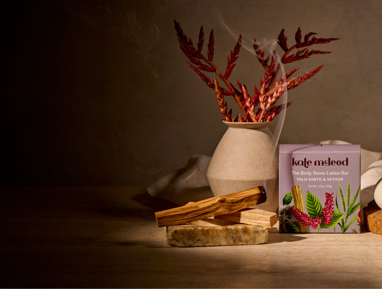 A styled product scene showing a box of Kafe Meleid body lotion bar next to a lit incense stick and dried botanicals in a ceramic vase, set on a wooden surface in warm, moody lighting.