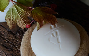 A round white body stone with an embossed Kate McLeod monogram, resting on a textured beige coaster or dish over a dark wooden surface, with green and brown leaves and small red berries placed on top.