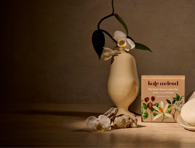 A styled product photo showing the kafe meleod Vanilla & Coffee body stone lotion bar box next to a beige ceramic vase with white orchids, on a wooden surface against a textured brown wall.