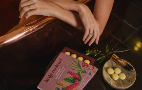 A person's hands resting on a dark table next to an open pink box of Kate McLeod Pebbles, displayed on a small wooden board and a copper tub in the background.