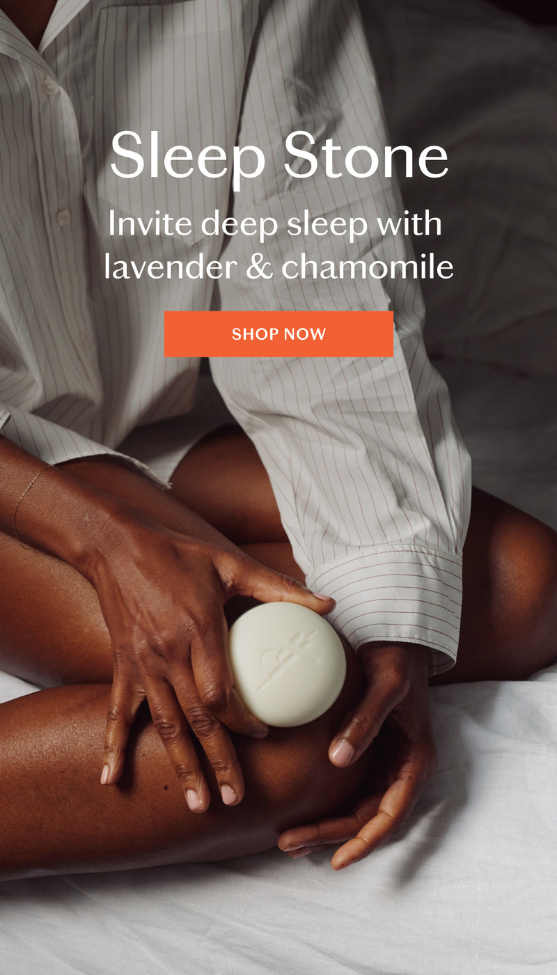 A person lying on a bed holds a round white soap bar labeled Sleep Stone over their knee. The person is wearing a white pinstriped shirt.