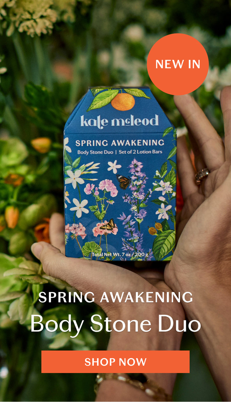 Hands holding Spring Body Stone Duo against plants in background with 'Spring Awakening Body Stone Duo' text and 'Shop Now' button. Click to shop Spring Body Stone Duo.