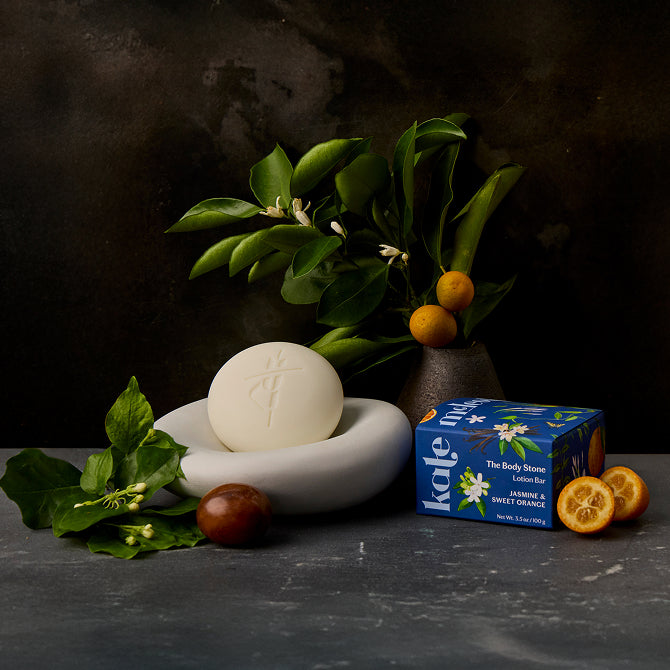 Jasmine & Sweet Orange Body Stone in stone dish with packaging, jasmine branches, and orange halves around.