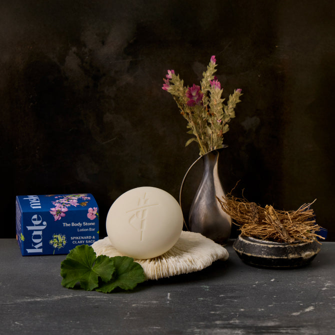 Spikenard & Clary Sage Body Stone lotion bar on a white plate with ingredients, spikenard and clary sage, arranged around it and packaging next to it.