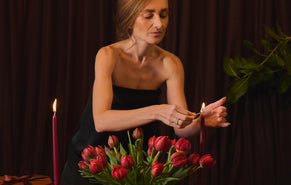 A woman in a black strapless dress lights a candle next to a bouquet of red tulips against a dark brown curtain background.
