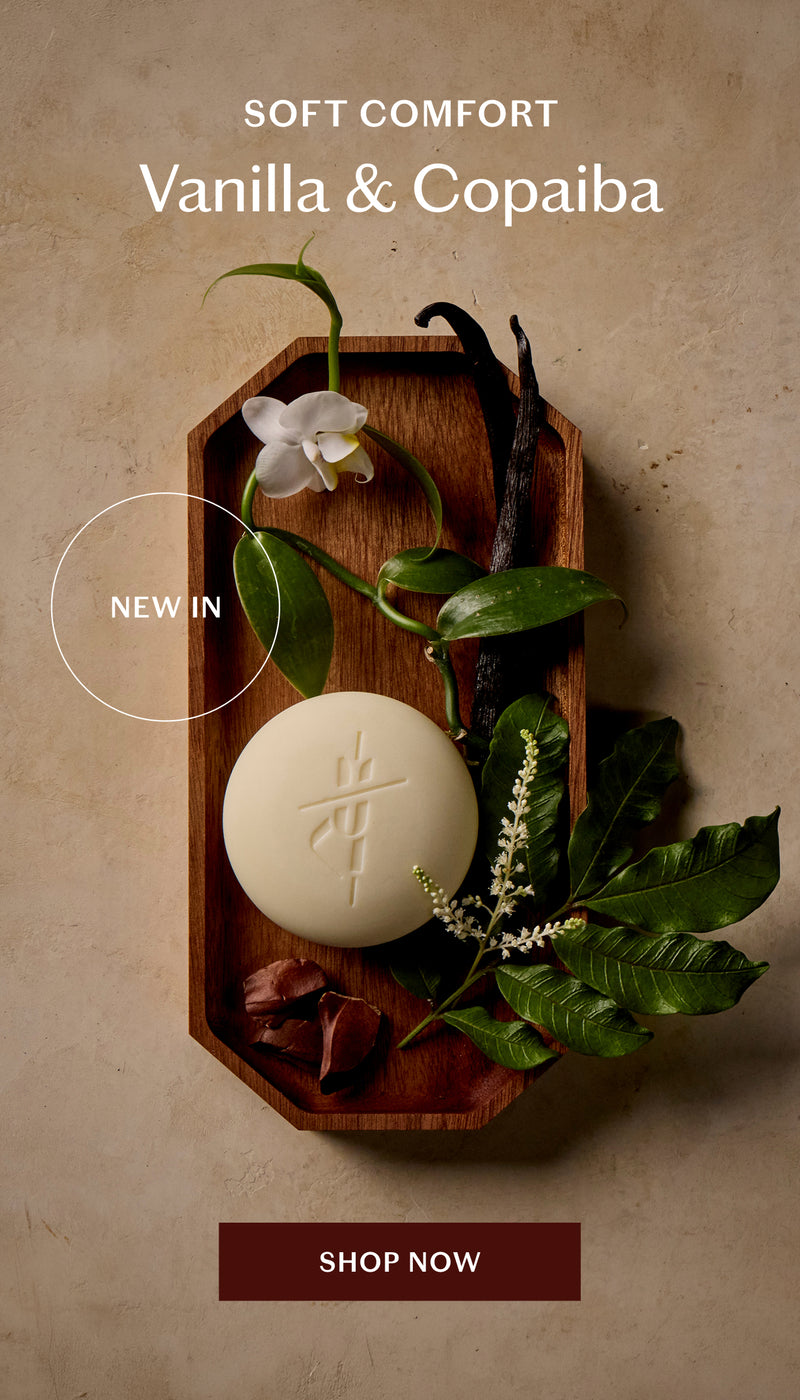A round cream-colored Body Stone lotion bar displayed on a wooden tray with green leaves, a white flower, and vanilla pods. The image includes text reading 'SOFT COMFORT Vanilla & Copaiba' and a 'SHOP NOW' button.